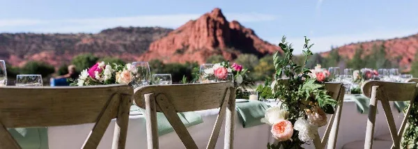 A wedding reception table setting featuring floral arrangements and wooden chairs, set against a backdrop of red rock desert mountains.