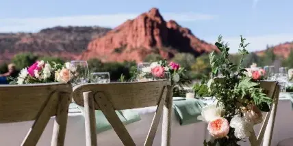 A wedding reception table setting featuring floral arrangements and wooden chairs, set against a backdrop of red rock desert mountains.