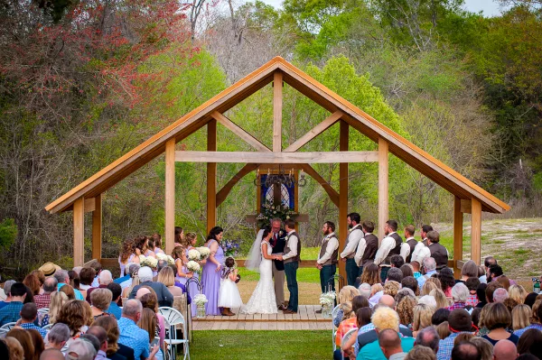 Outdoor wedding ceremony at a wooden chapel with a bride, groom, and guests in a wooded setting.