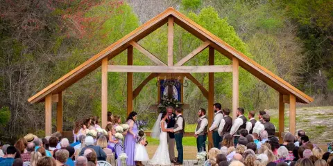 Outdoor wedding ceremony at a wooden chapel with a bride, groom, and guests in a wooded setting.