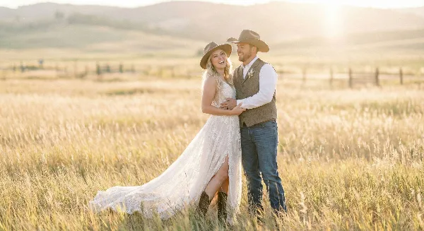 A bride and groom in cowboy hats embracing in a golden sunlit field, bride in a lace gown, groom in a vest and jeans.