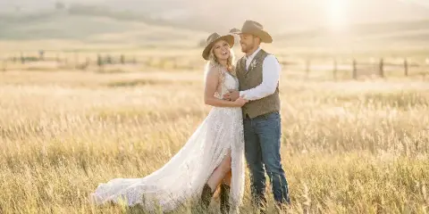 A bride and groom in cowboy hats embracing in a golden sunlit field, bride in a lace gown, groom in a vest and jeans.