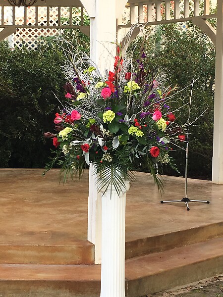 A large, colorful floral arrangement featuring red, purple, and yellow flowers on a white pedestal in an outdoor setting.