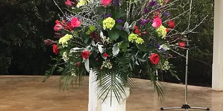 A large, colorful floral arrangement featuring red, purple, and yellow flowers on a white pedestal in an outdoor setting.