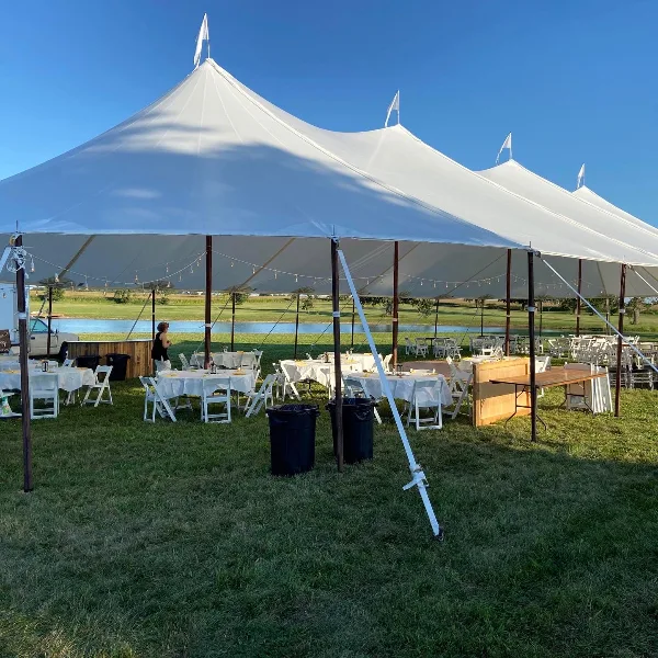 Outdoor event setup featuring a large white tent with tables, chairs, and linens arranged on a grassy field.