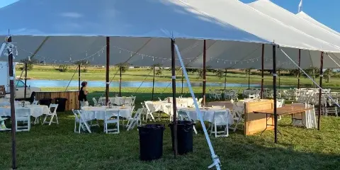 Outdoor event setup featuring a large white tent with tables, chairs, and linens arranged on a grassy field.