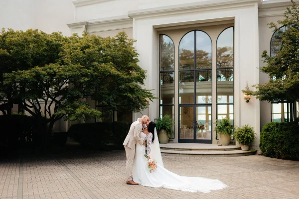 A bride and groom share a kiss in front of an elegant white building with arched windows and greenery.
