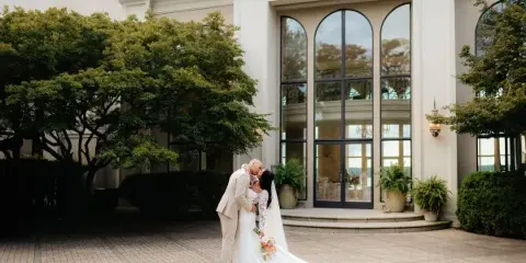 A bride and groom share a kiss in front of an elegant white building with arched windows and greenery.