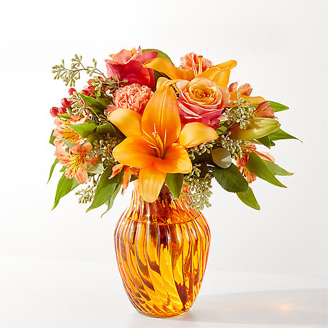 A vibrant floral arrangement featuring orange lilies, roses, and alstroemeria in a textured amber glass vase.