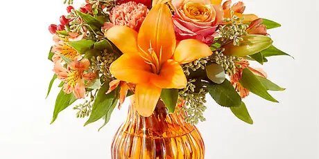 A vibrant floral arrangement featuring orange lilies, roses, and alstroemeria in a textured amber glass vase.