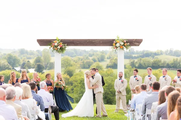 Outdoor wedding ceremony with bride and groom kissing under a floral arch, surrounded by wedding party and guests in a grassy field with rolling hills in the background.