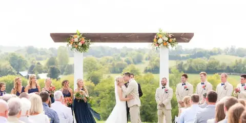 Outdoor wedding ceremony with bride and groom kissing under a floral arch, surrounded by wedding party and guests in a grassy field with rolling hills in the background.