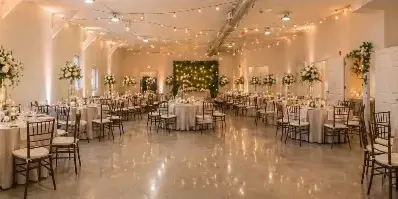 Interior of a wedding venue with round tables, white linens, floral centerpieces, and string lights.