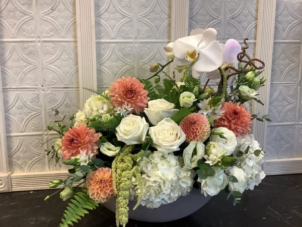 A custom floral arrangement featuring white roses, peach dahlias, and white orchids in a grey ceramic vessel.