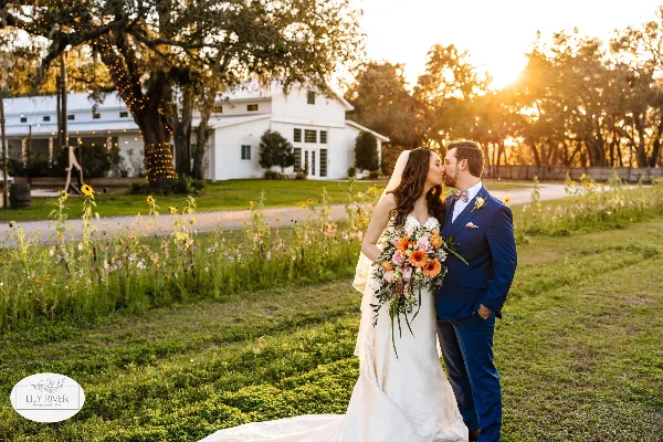 A bride and groom kissing in a field with a white barn and string lights in the background at sunset.