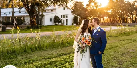 A bride and groom kissing in a field with a white barn and string lights in the background at sunset.