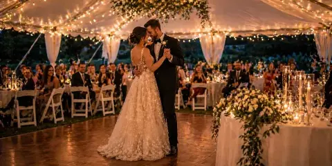 A bride and groom dancing at a wedding reception under a large tent with string lights, surrounded by tables and white chairs.
