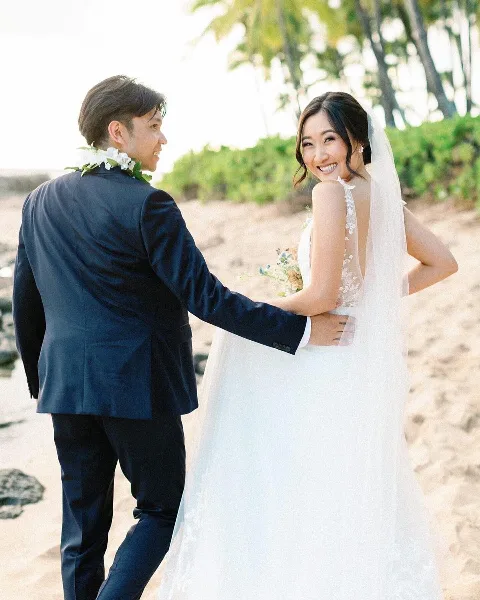 A bride and groom standing on a sandy beach with palm trees in the background, the bride wearing a white wedding dress and veil.