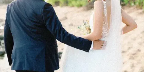A bride and groom standing on a sandy beach with palm trees in the background, the bride wearing a white wedding dress and veil.
