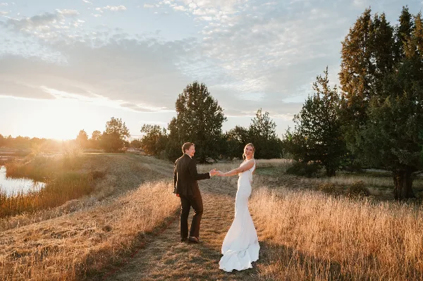 A bride and groom holding hands and smiling at each other in a grassy field at sunset.