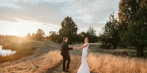 A bride and groom holding hands and smiling at each other in a grassy field at sunset.