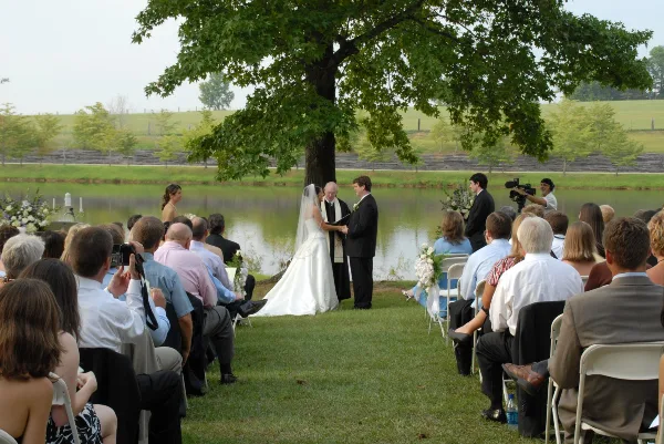A wedding ceremony in progress outdoors near a lake with guests seated in chairs.