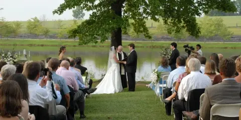 A wedding ceremony in progress outdoors near a lake with guests seated in chairs.