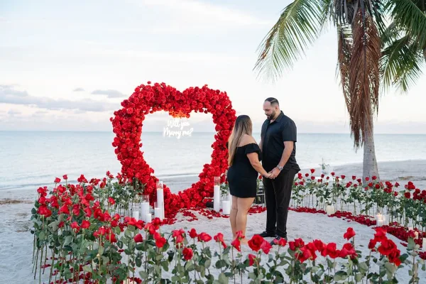 A couple standing on a sandy beach in front of a large red rose heart arch with a 'Will you Marry me?' sign, surrounded by red roses and white candles.