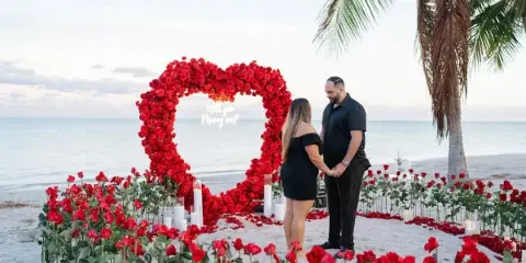 A couple standing on a sandy beach in front of a large red rose heart arch with a 'Will you Marry me?' sign, surrounded by red roses and white candles.
