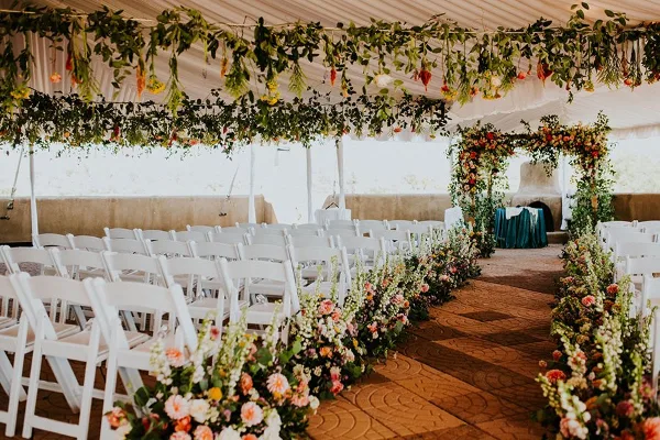 Interior of a white event tent featuring a floral arch, hanging greenery, and rows of white chairs set up for a wedding ceremony.