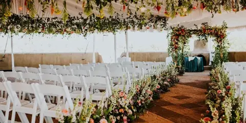 Interior of a white event tent featuring a floral arch, hanging greenery, and rows of white chairs set up for a wedding ceremony.