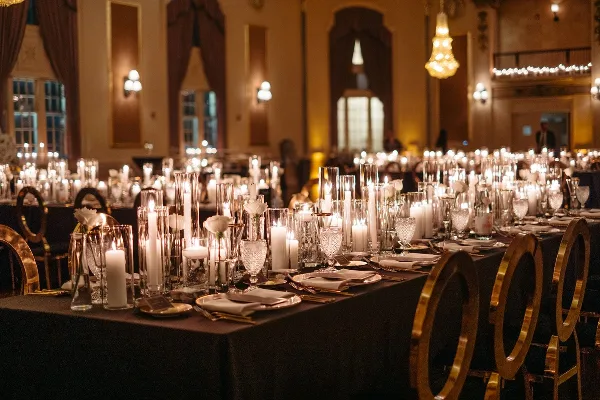 A formal wedding reception table setting featuring tall glass candle holders, white roses, and gold-rimmed chairs in a grand ballroom.