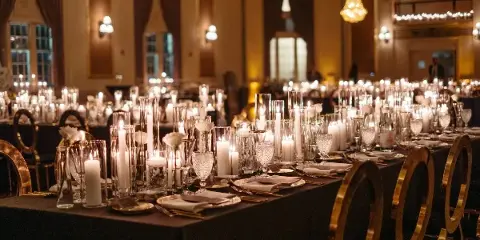 A formal wedding reception table setting featuring tall glass candle holders, white roses, and gold-rimmed chairs in a grand ballroom.