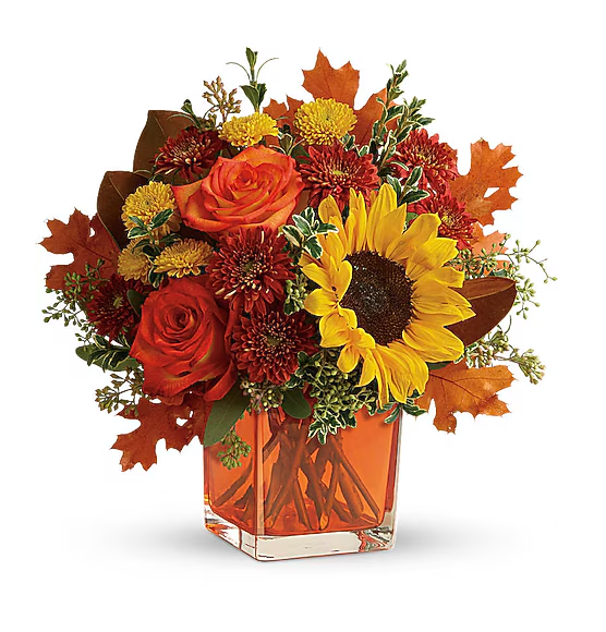 A floral arrangement with red roses, a yellow sunflower, and autumn leaves in a square glass vase.