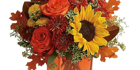 A floral arrangement with red roses, a yellow sunflower, and autumn leaves in a square glass vase.