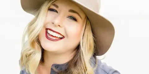 Portrait of a smiling blonde woman wearing a beige hat and a grey shirt.
