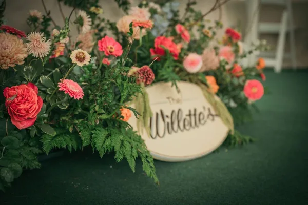 A close-up of a vibrant, garden-style floral arrangement featuring pink and peach roses, dahlias, and lush greenery, set against a dark green background with a decorative sign.
