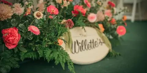 A close-up of a vibrant, garden-style floral arrangement featuring pink and peach roses, dahlias, and lush greenery, set against a dark green background with a decorative sign.