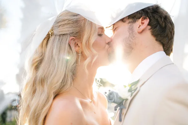 A bride and groom sharing a kiss under a white canopy with bright sunlight in the background.