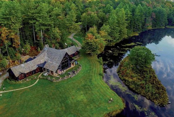 Aerial view of the Lakefalls estate featuring a rustic stone and timber lodge situated on a grassy lawn next to a lake, surrounded by dense forest.