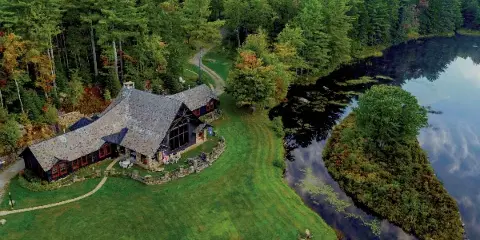 Aerial view of the Lakefalls estate featuring a rustic stone and timber lodge situated on a grassy lawn next to a lake, surrounded by dense forest.
