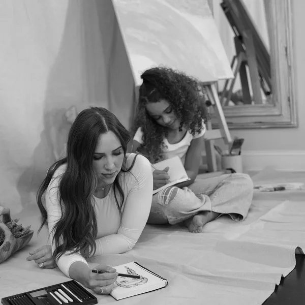 A black and white photograph of a woman with long dark hair drawing in a sketchbook while a young girl sits behind her, also drawing, in a creative studio space.