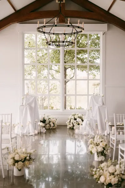Interior of a wedding venue with a round chandelier, large windows, white floral arrangements, and a polished floor.