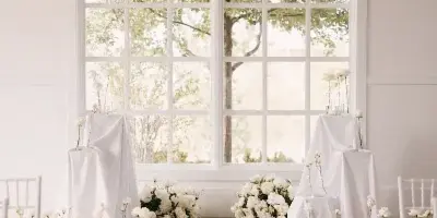 Interior of a wedding venue with a round chandelier, large windows, white floral arrangements, and a polished floor.