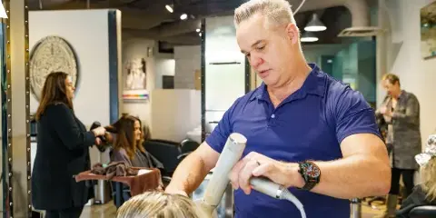 A male hair stylist in a blue polo shirt blow-drying a client's hair in a busy salon.
