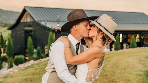 A bride and groom kissing in front of a rustic barn with a mountain view.