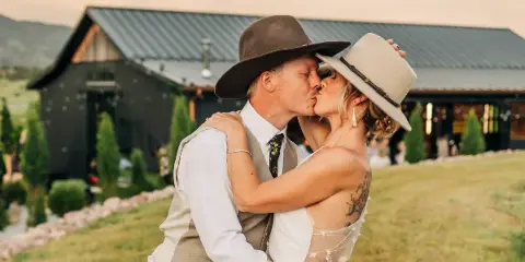 A bride and groom kissing in front of a rustic barn with a mountain view.