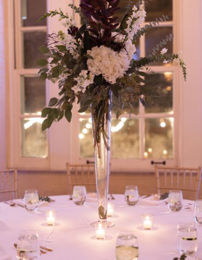 A tall, elegant floral arrangement with white and purple flowers in a glass vase, placed on a white tablecloth with candles.