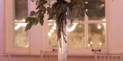 A tall, elegant floral arrangement with white and purple flowers in a glass vase, placed on a white tablecloth with candles.