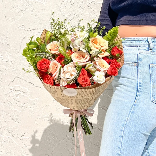 A hand-tied bouquet of red and peach roses wrapped in newspaper-style paper with a Fliorens ribbon.
